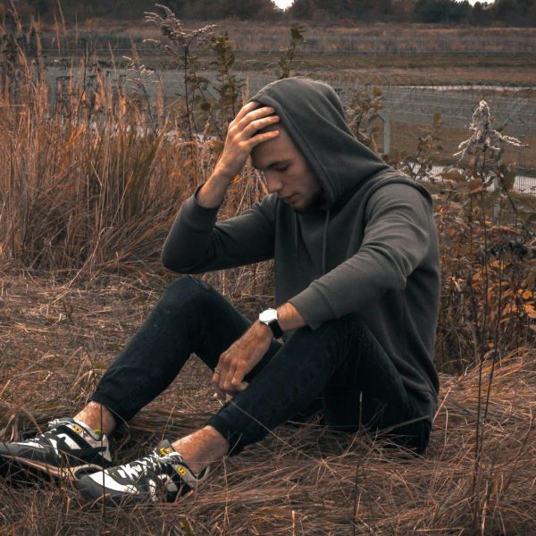Hombre con sudadera sentado en un suelo con vegetación, con gesto preocupado echándose una mano en la frente